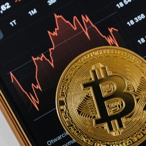 A close-up of a gold Bitcoin coin placed on a smartphone displaying a stock market chart.